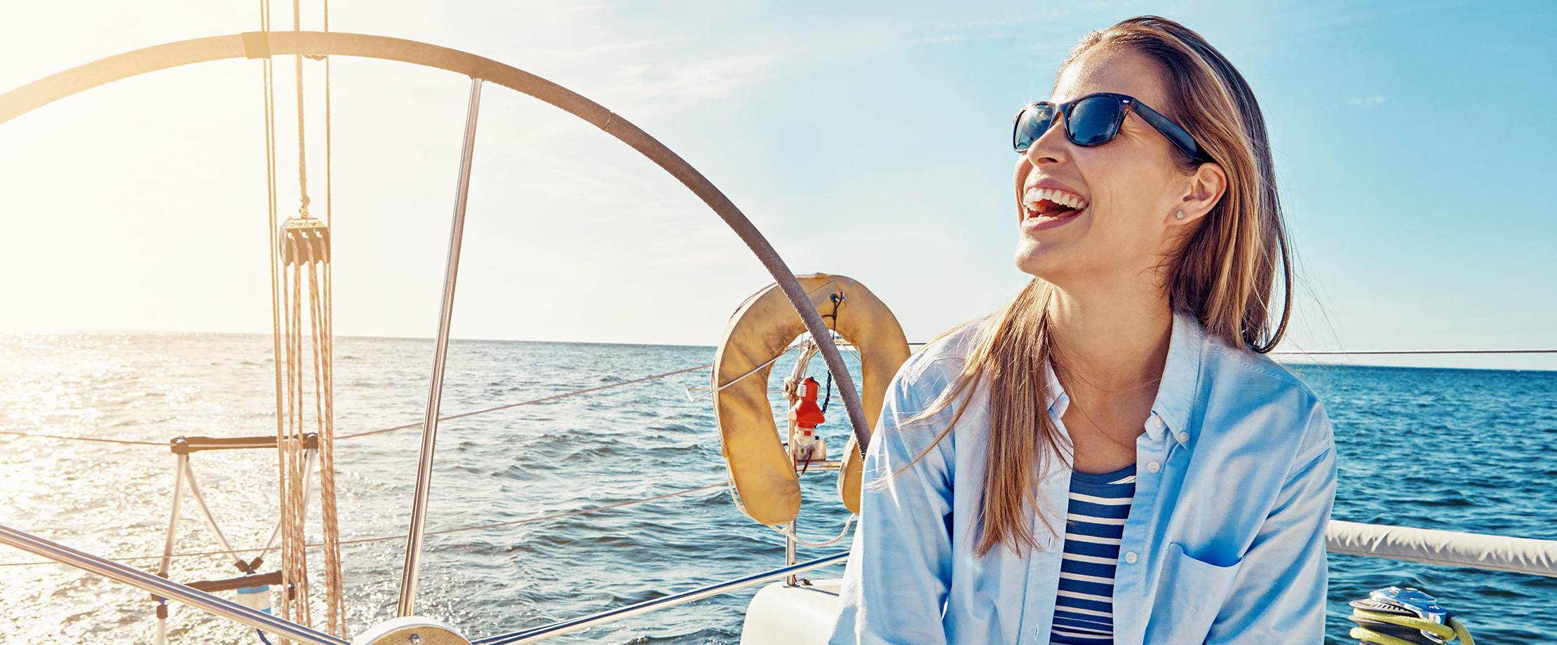 Eine blonde Frau sitzt auf einem Segelboot und schaut lachend in den Himmel.