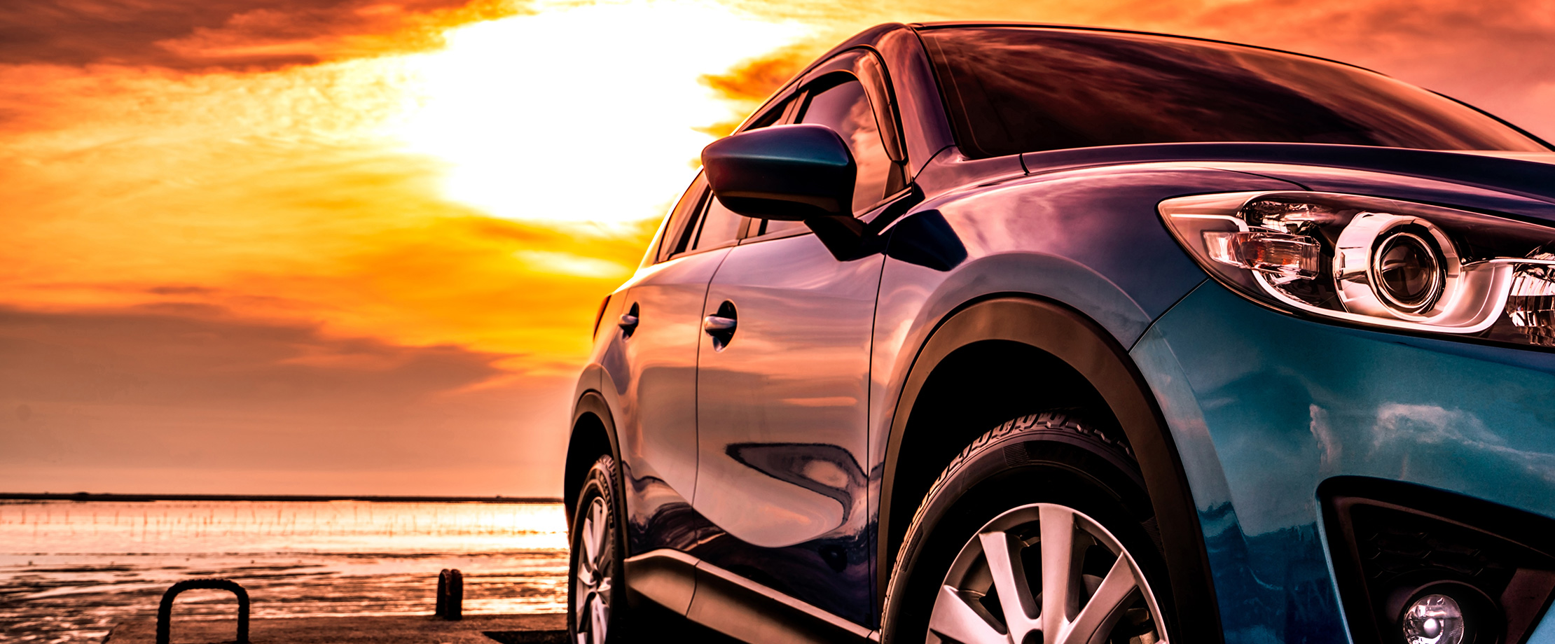 Ein blaues Auto steht bei Sonnenuntergang am Meer. Man sieht die rechte Seite des Autos, in der sich der gelb-orange Himmel und der Strand spiegelt.