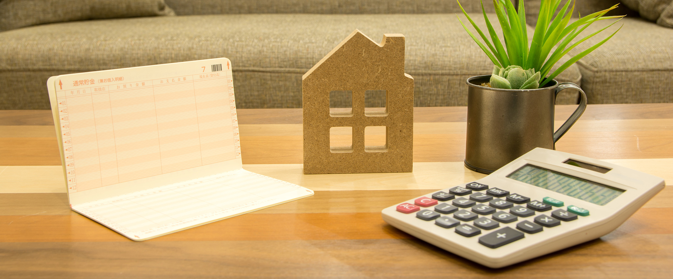 Auf einem Tisch sind ein Taschenrechner, ein Finanzbuch und ein Holzmodell eines Hauses platziert. 