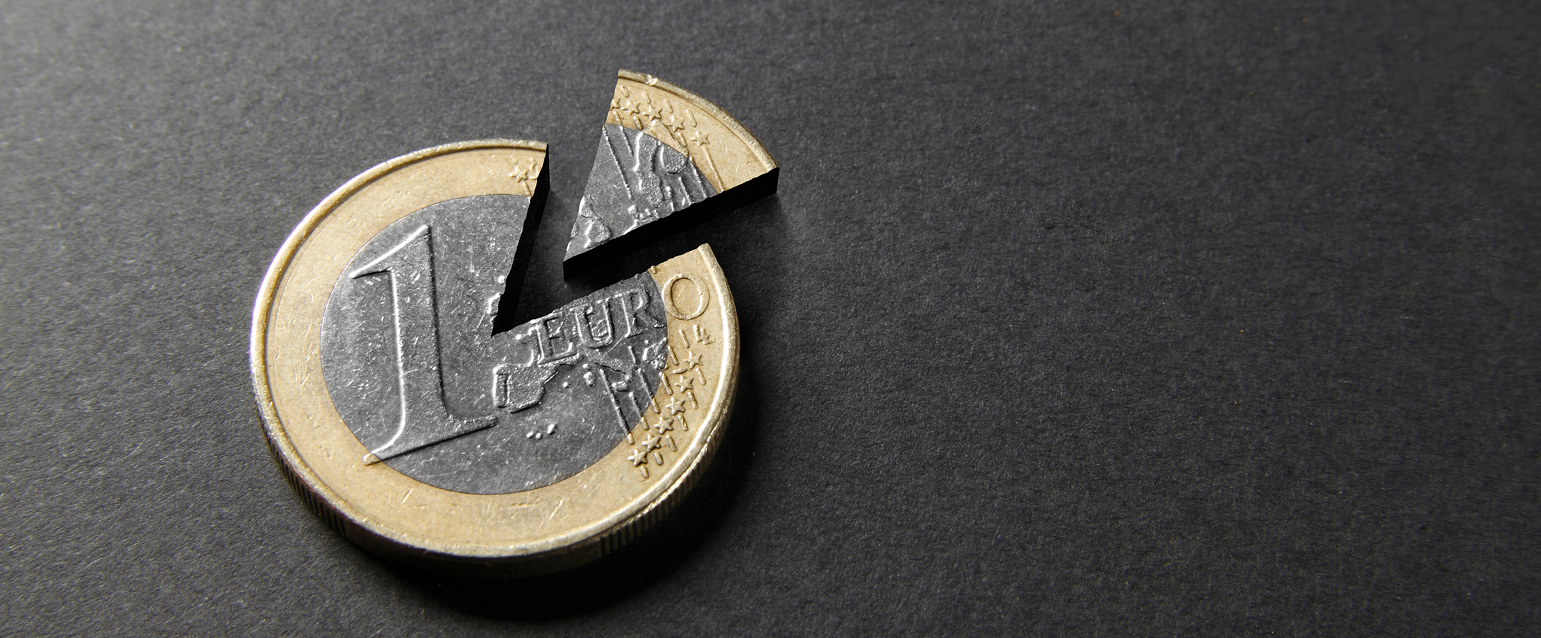 Ein 1-Euro-Sück liegt auf einer schwarzen Tischplatte. Ein kuchenförmiges Stück wurde aus dem Euro ausgeschnitten und liegt abgetrennt daneben. 