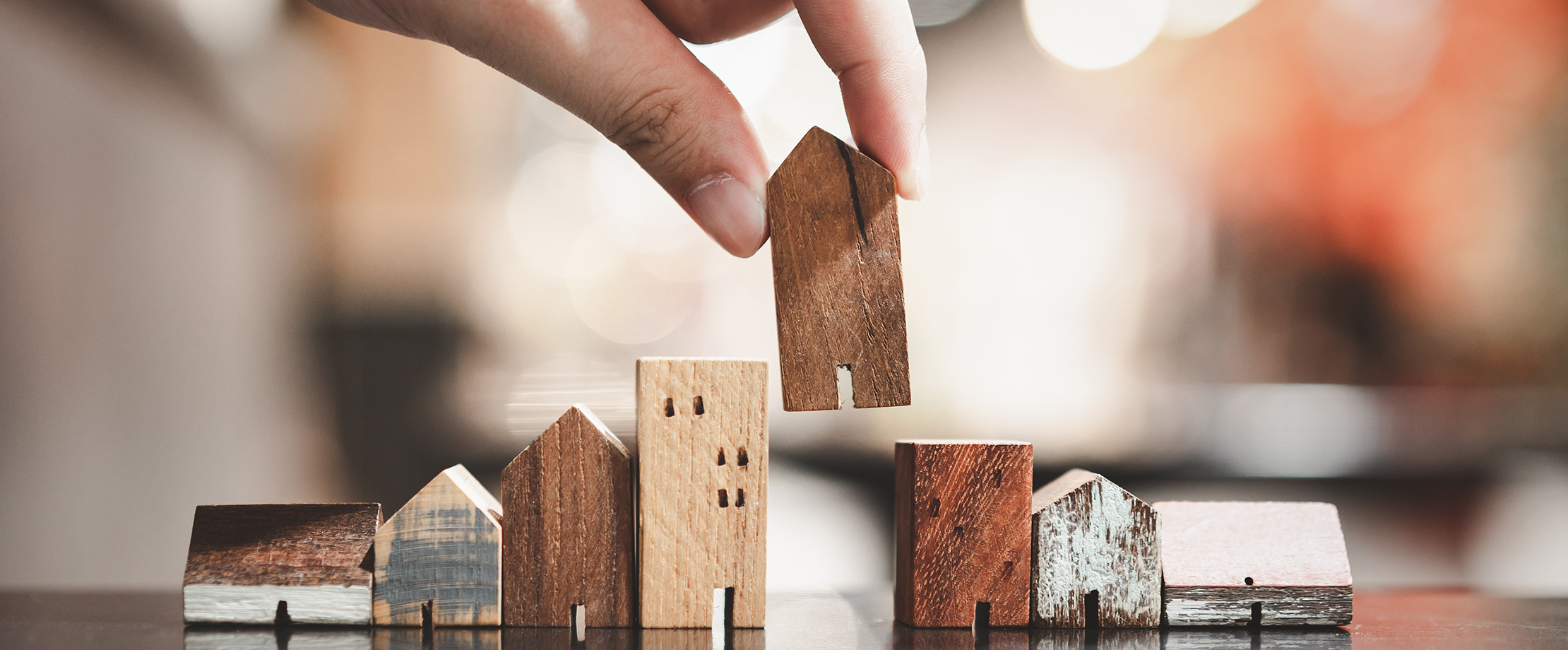 Kleine Holzhäuser stehen in einer Reihe. Von oben wird von einer Hand ein weiteres Holzhaus in die Reihe eingesetzt. 