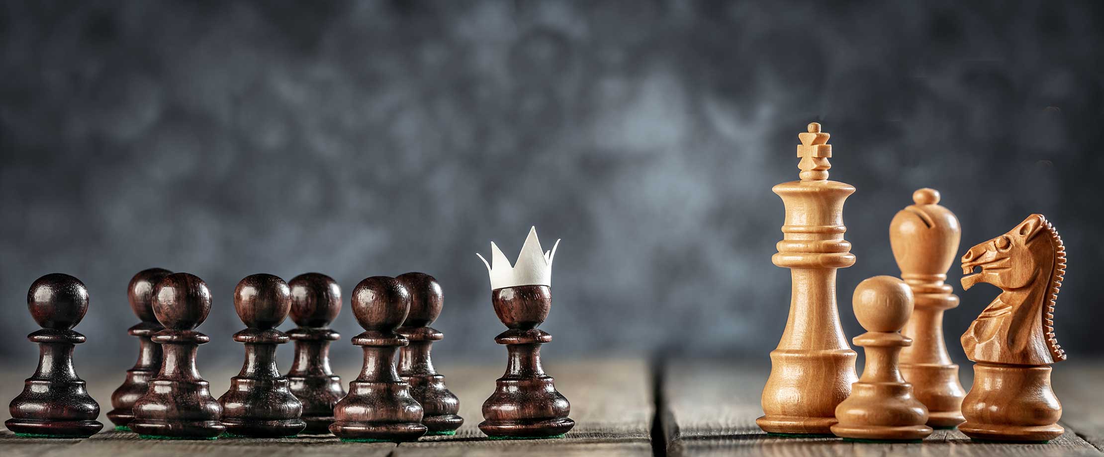 Schachfiguren stehen auf einem Tisch vor schwarzem Hintergrund. Die weißen Figuren rechts sind der König, ein Läufer, Ein Pferd und ein Bauer. Die schwarzen Figuren links sind alle Bauern, der vorderste von ihnen trägt eine Papierkrone.