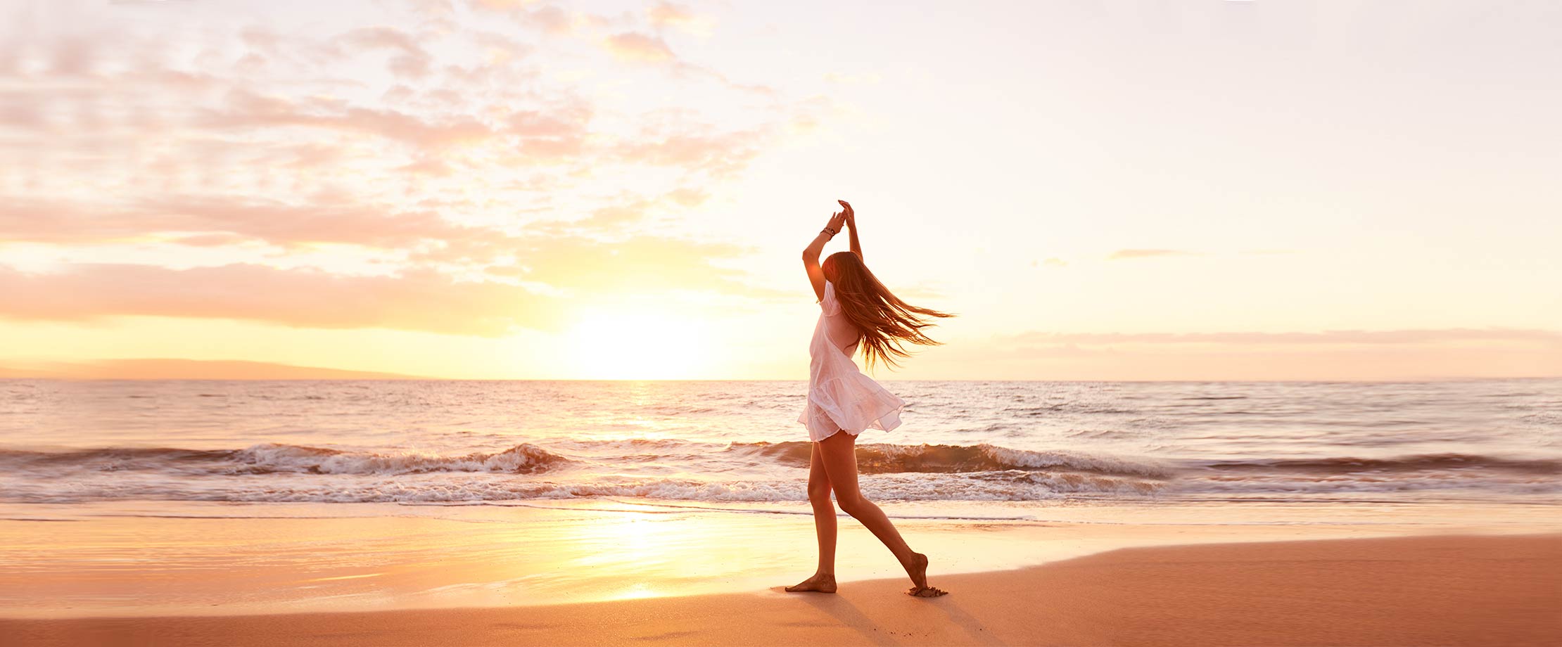 Eine junge Frau mit langen, hellen Haaren tanzt in einem weißen, kurzen Sommerkleid den Strand entlang. Sie hat die Hände während einer Drehung erhoben. Im Hintergrund sieht man den Sonnenuntergang über dem Meer.