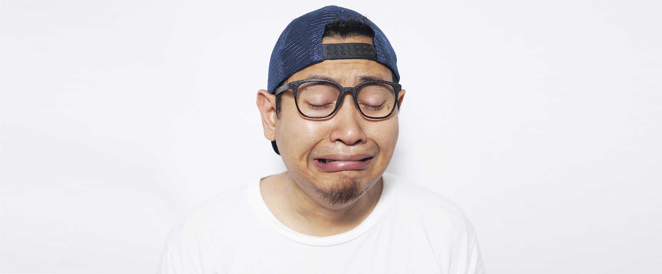 Ein Mann mit Brille, blauer, verkehrt aufgesetzter Kappe und weißem T-Shirt steht vor einer weißen Wand und weint. Sein Gesicht ist vor Trauer verzogen.