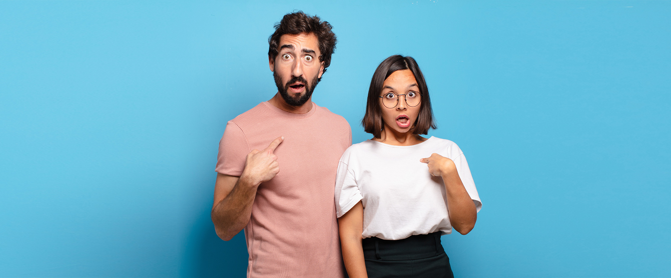 Ein Mann und eine Frau stehen vor einem hellblauen Hintergrund und haben geschockt die Augen aufgerissen. Mit ihren Fingern deuten sie mit einer 