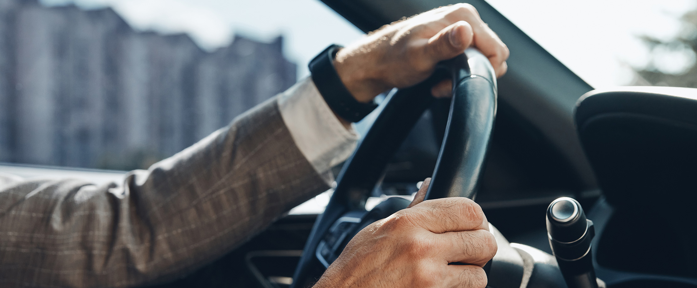 Ein Mann im Anzug fährt mit dem Auto. Man kann seine Arme und Hände sehen, mit denen er das Lenkrad hält. Am Handgelenk trägt er eine schwarze Armbanduhr.