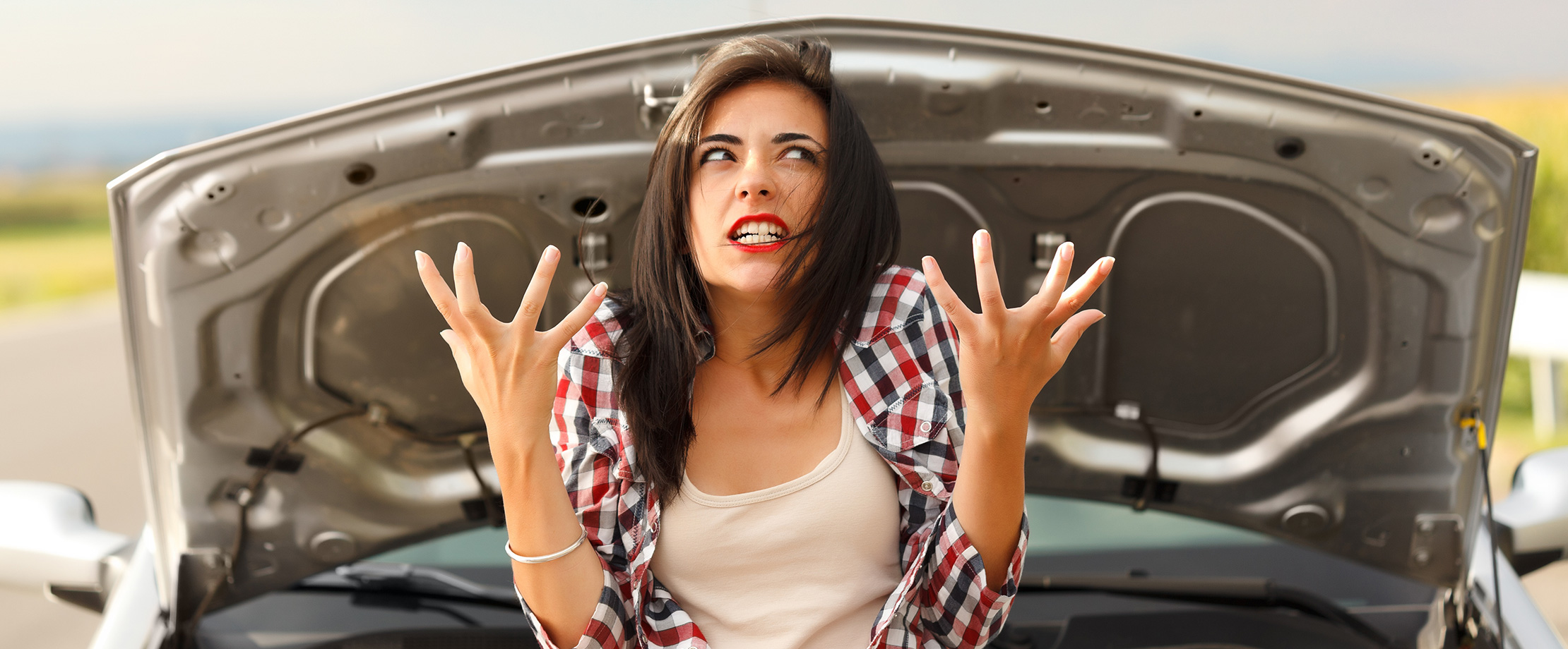 Eine schwarzhaarige Frau sitzt vor der geöffneten Motorhaube ihres Autos. Ihr Gesicht ist genervt verzogen und sie hat die Hände in einer verzweifelten Geste erhoben.