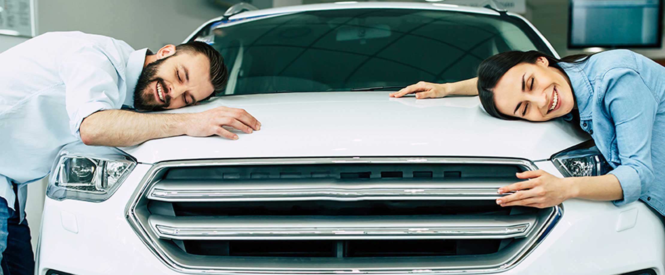 Ein junges paar liegt lächelnd auf der Motorhabe ihres neuen weißen Autos. Sie haben die Augen geschlossen. 
