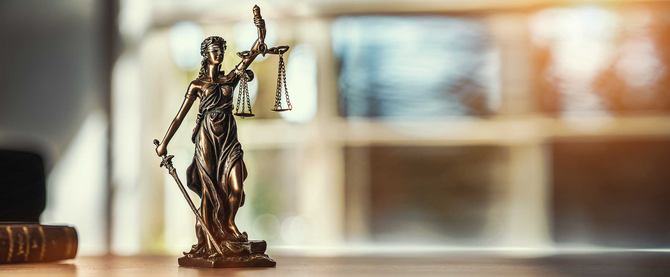 Eine Statuette der Göttin der Gerechtigkeit steht auf einem Tisch. In der einen Hand hält sie eine Waage, in der anderen ein Schwert. Daneben liegt ein Richterhammer. 
