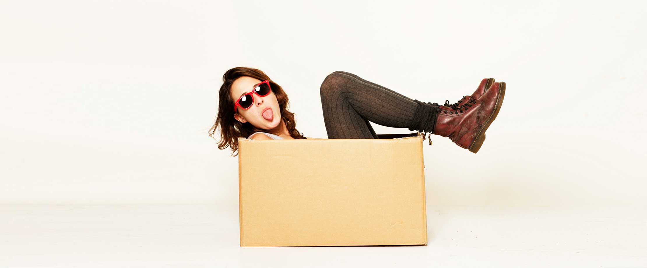 Eine junge Frau mit Sonnenbrille streckt frech ihre Zunge heraus. Sie sitzt in einem Karton und hält ihre Füße angewinkelt in die Luft.
