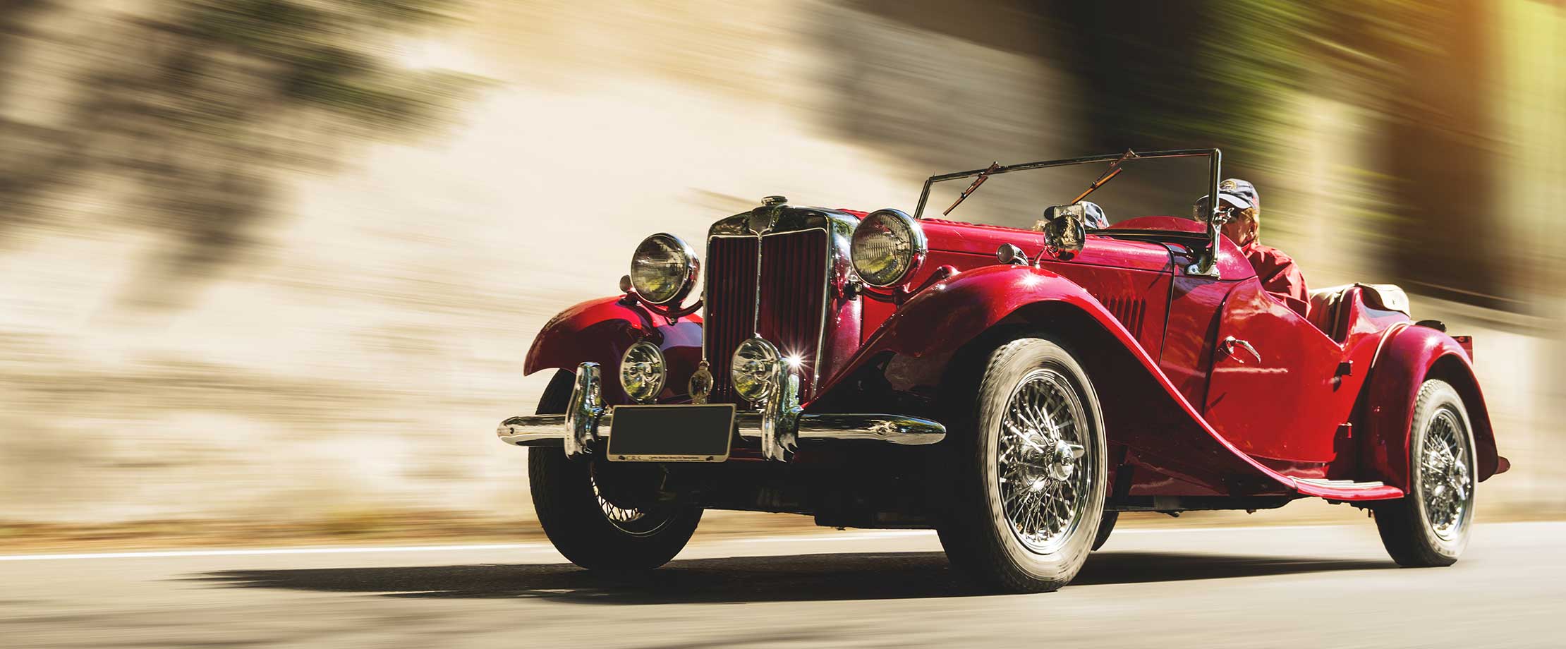 Ein roter Oldtimer fährt eine Straße entlang. Der Hintergrund ist aufgrund der schnellen Fahrt verschwommen.