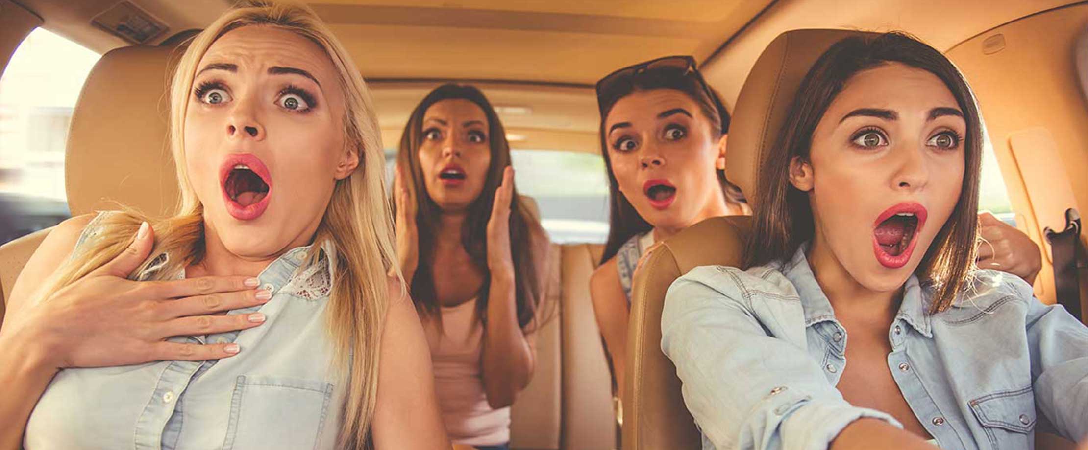Vier Freundinnen sitzen in einem Auto und schauen erschrocken nach vorne. Sie haben den Mund und die Augen vor Entsetzen weit aufgerissen.