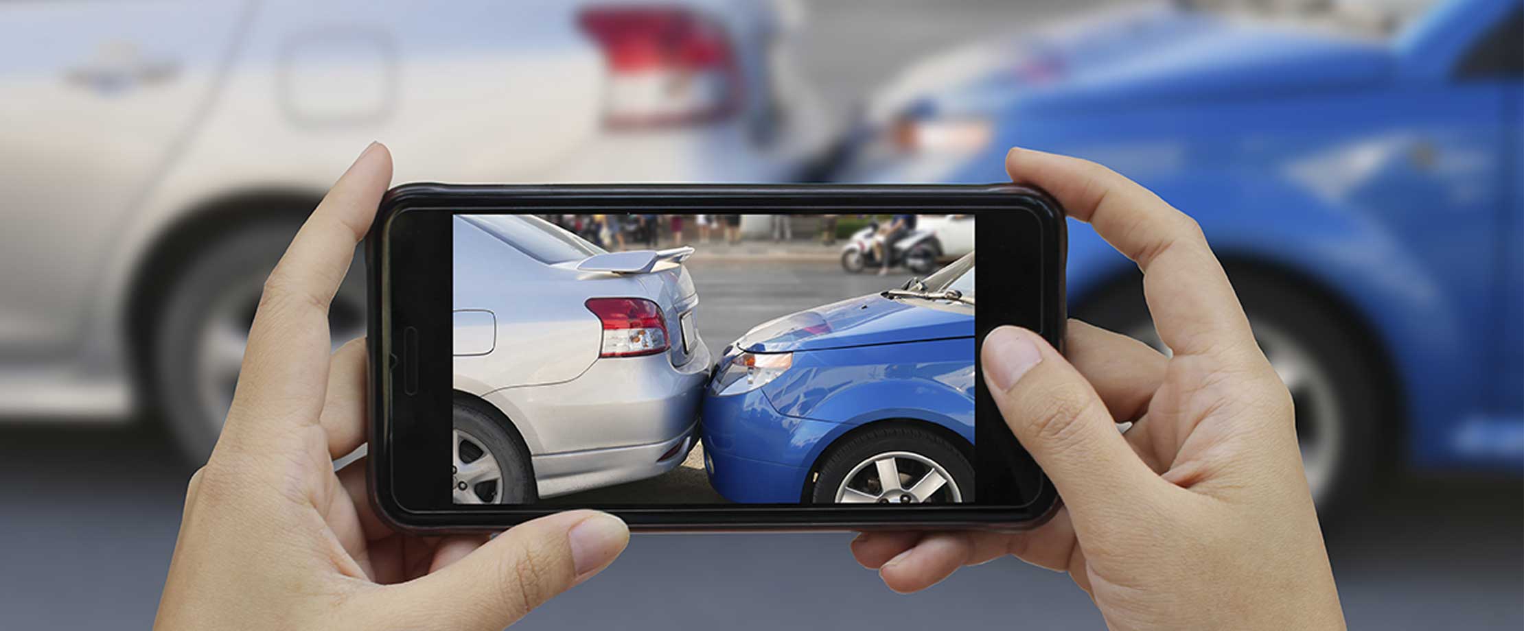 Zwei Hände halten ein Handy um ein Foto von einem Unfall zu machen. Ein blaues Auto ist auf das Heck eines silbernen Autos aufgefahren.