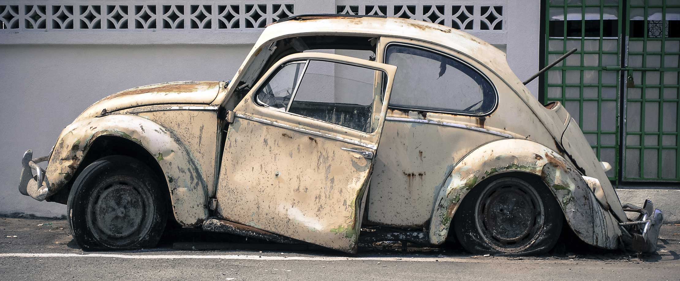 Ein alter VW Käfer steht komplett zerstört vor einem weißen Gebäude. Die Tür hängt nur noch an wenigen Schrauben, das Heck liegt auf dem Boden auf.