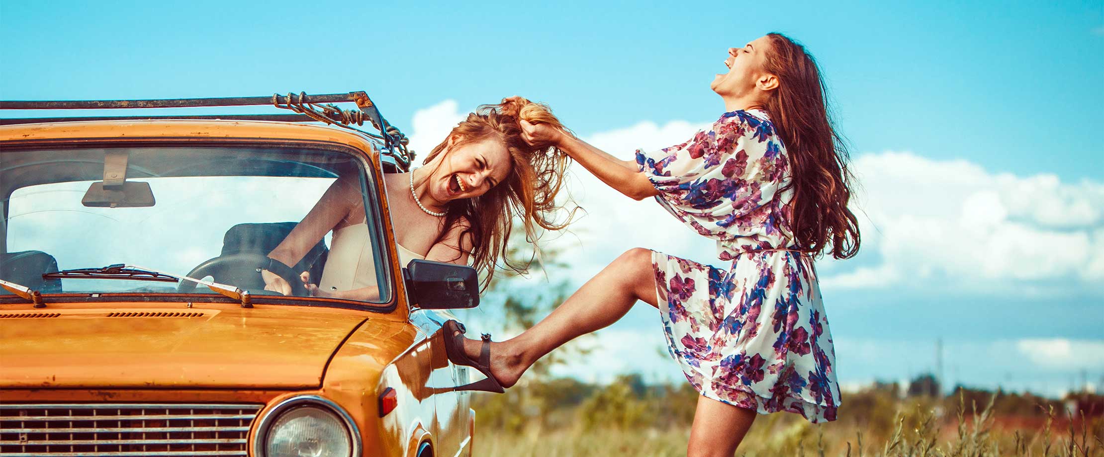 Eine junge Frau im Sommerkleid zerrt an den Haaren einer anderen Frau, die in einem gelben Auto sitzt. Die beiden haben die Augen geschlossen und den Mund zum Schreien geöffnet.