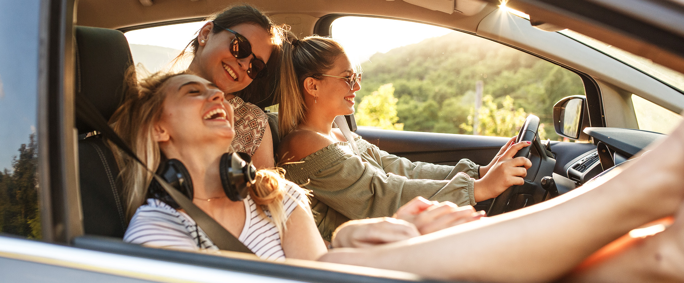 Drei Freundinnen sitzen im Sommer lachend in einem Auto und fahren in einer grünen Landschaft.