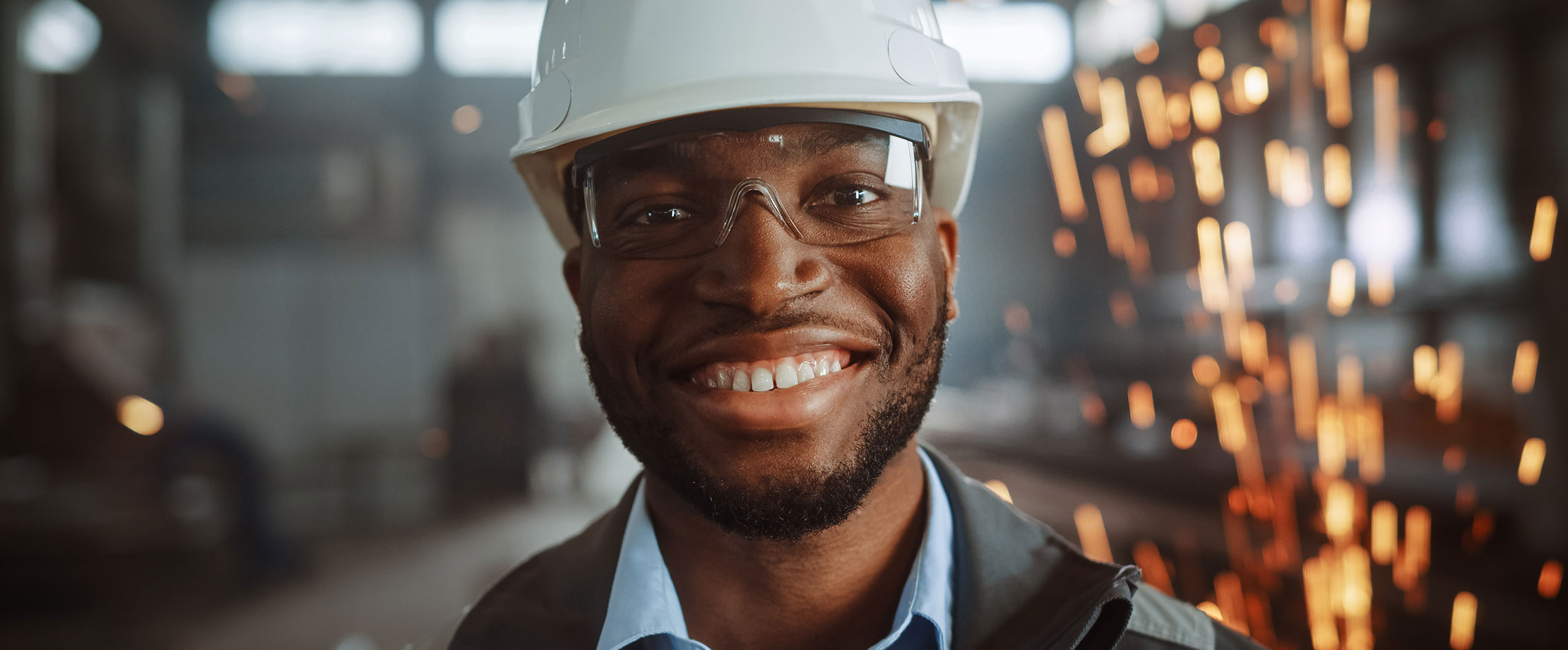 Ein Mann steht in einer Werkstatt und lächelt in die Kamera, er trägt einen Schutzhelm und eine Schutzbrille, im Hintergrund fliegen Funken. 