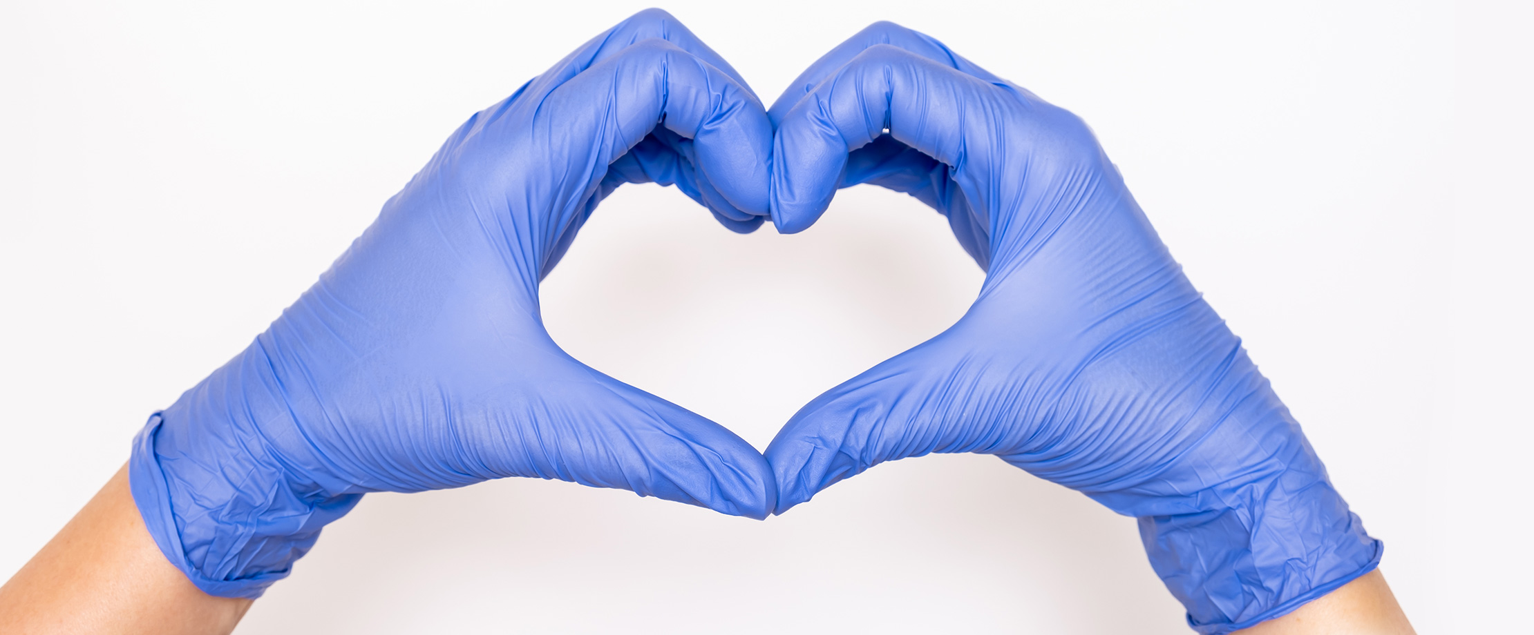Zwei Hände in blauen Chirurgenhandschuhen formen das Love-Sign. Dieses blaue, behandschuhte Herz ist vor einem sterilen weißen Hintergrund zu sehen.