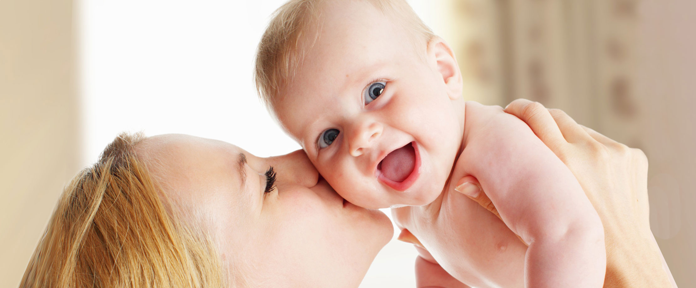 Eine Mutter hält ihr lachendes Baby in die Höhe und küsst es auf die Wange. Das Gesicht des Babys ist der Kamera zugewandt. 