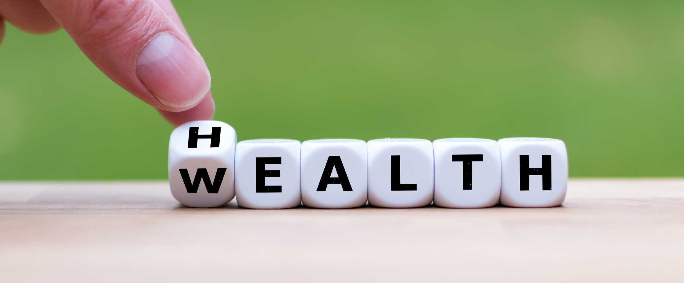 Auf einem Tisch vor einem grünen Hintergrund liegen Würfel, auf denen Buchstaben abgebildet sind. Daraus ergeben die Wörter „Wealth“ und „Health“.