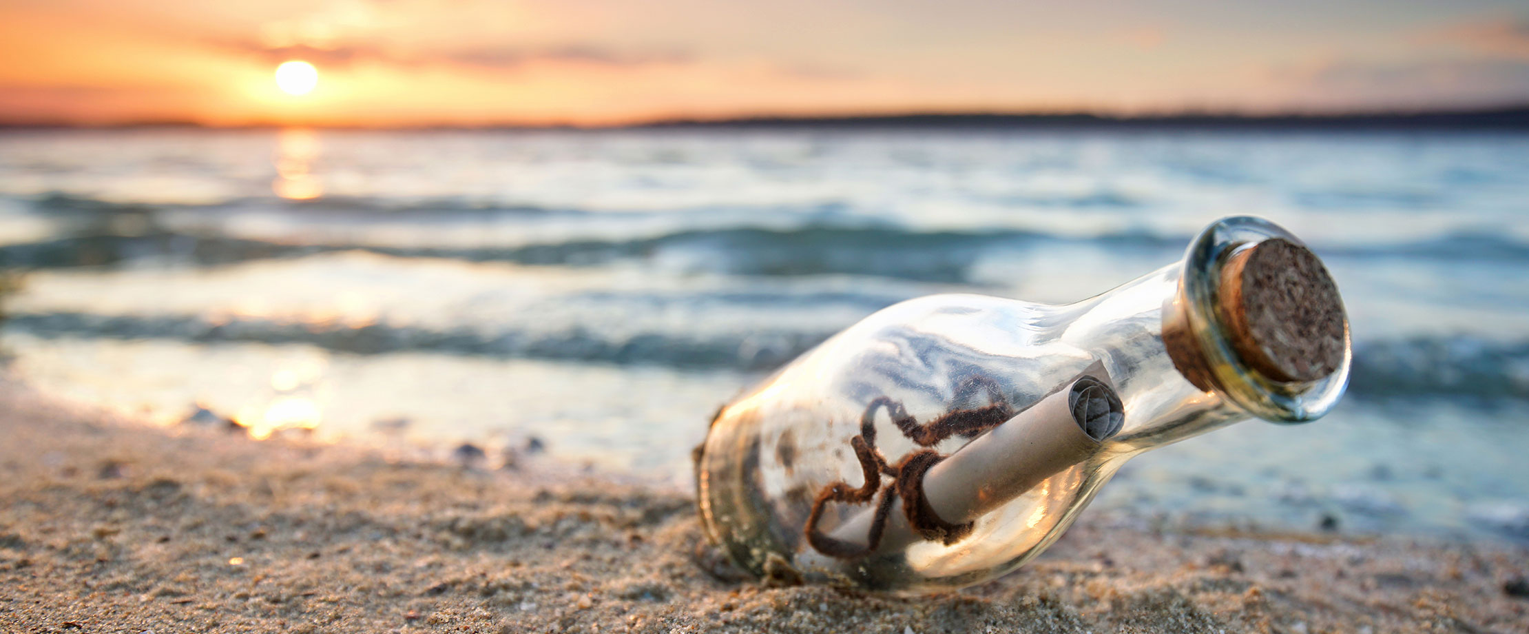 Eine Flaschenpost wurde am Strand angeschwemmt und liegt im Sand. Im Hintergrund geht die Sonne hinter dem Meer unter. Der Wellengang ist sanft.