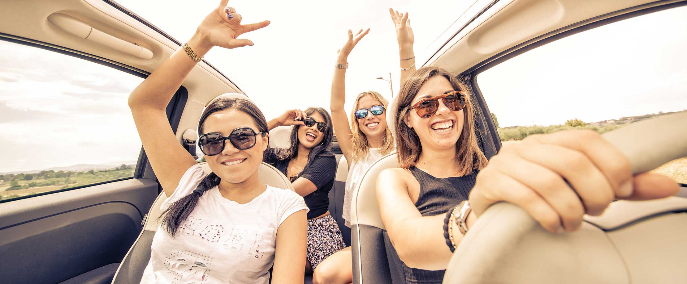 Vier Freundinnen sind zusammen auf einem Roadtrip und sitzen im Auto. Sie lachen in die Kamera und haben jubelnd die Hände in der Luft.