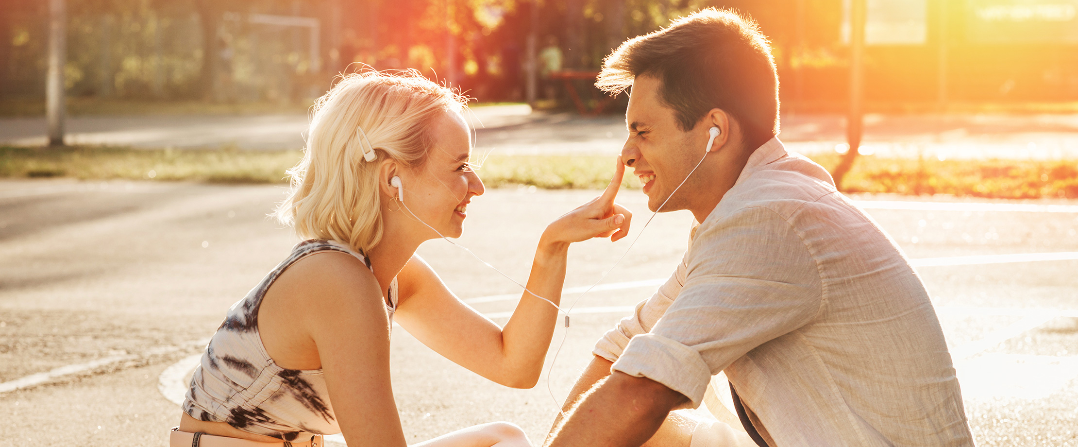 Ein junges Paar sitzt in der Abendsonne und lacht sich an, während sie sich Kopfhörer teilen und Musik hören. Die Frau hält scherzhaft ihren Finger auf die Nase des Mannes.
