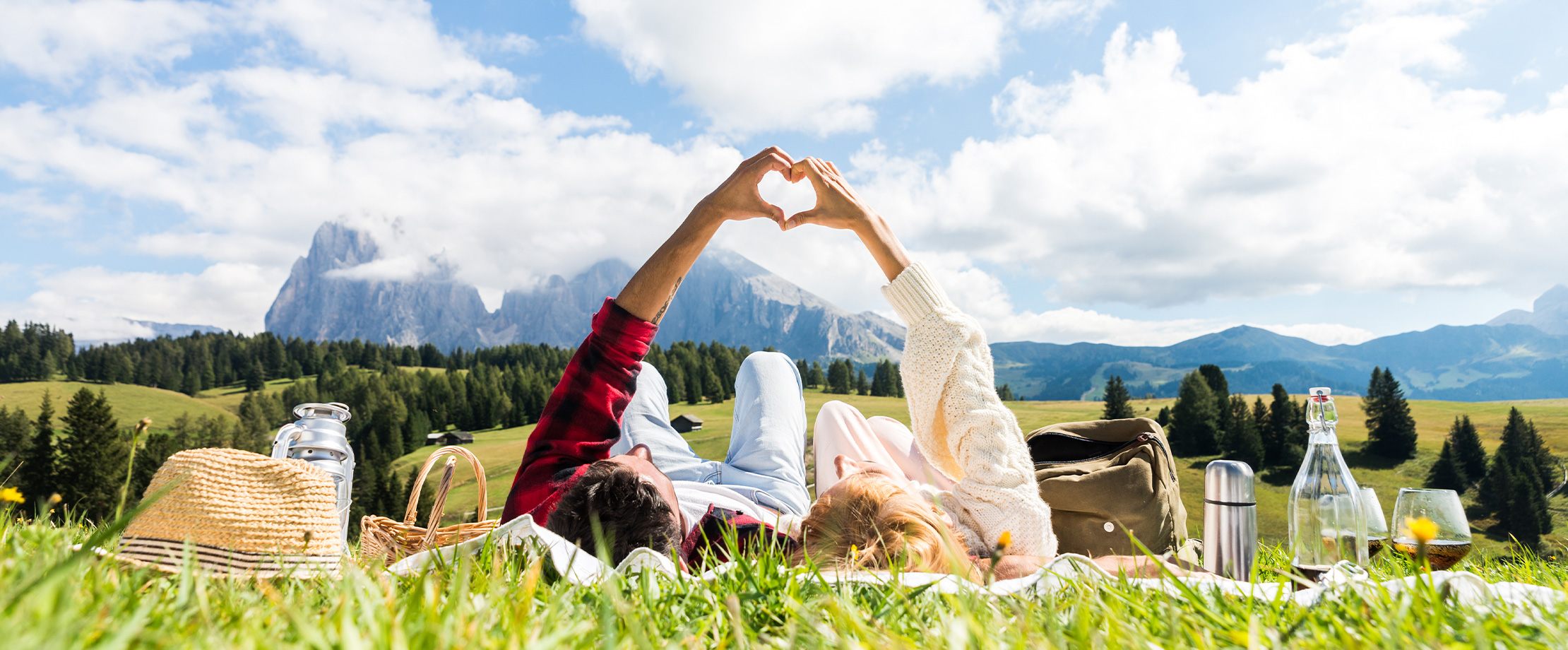 Pärchen liegt auf einer Picknickdecke auf der Alm. Mit den Händen formen sie ein Herz. Man kann das Bergpanorama und einen blauen Himmel mit Wolken sehen.