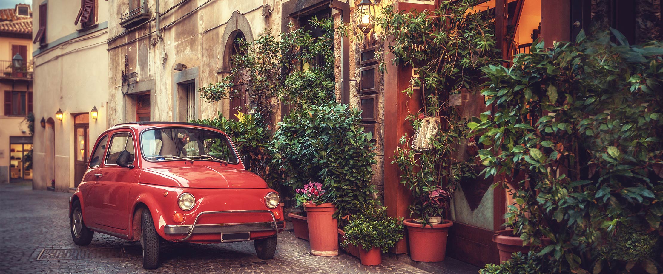 Ein rotes Auto steht vor einem Haus in einer schmalen Seitengasse Italiens. Vor dem Haus stehen viele Pflanzen, Efeu rankt sich die Hausmauer hinauf.