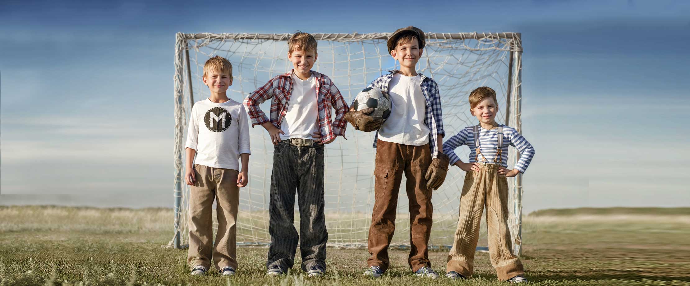 Vier Buben stehen lächelnd im Freien vor einem Tor. Sie tragen Cordhosen und karierte Hemden. Der größte von ihnen trägt Torwarthandschuhe und hält einen Fußball.