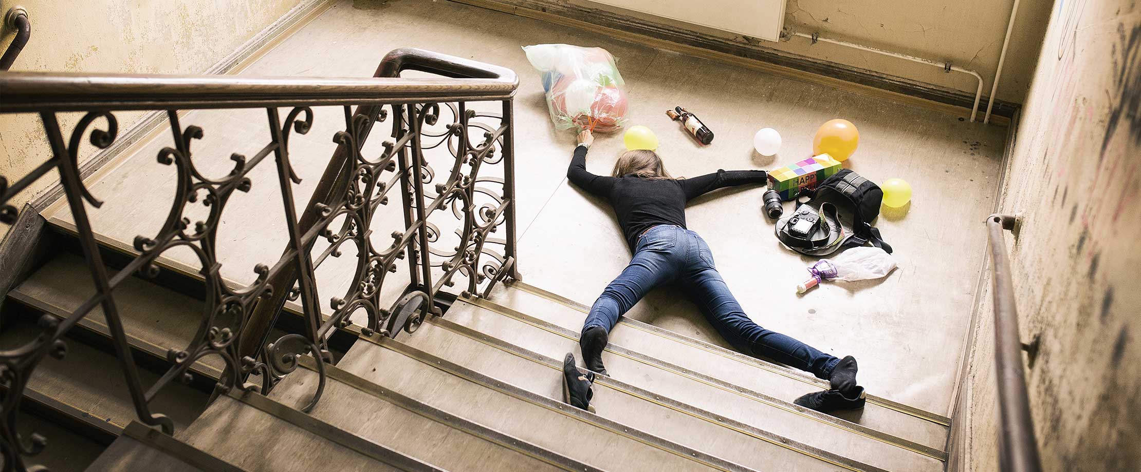 Eine Frau liegt am Fuß einer Treppe, nachdem sie gestürzt ist. Um Sie herum liegen Einkäufe, ihre Tasche, eine Kamera und ein Geburtstagsgeschenk.
