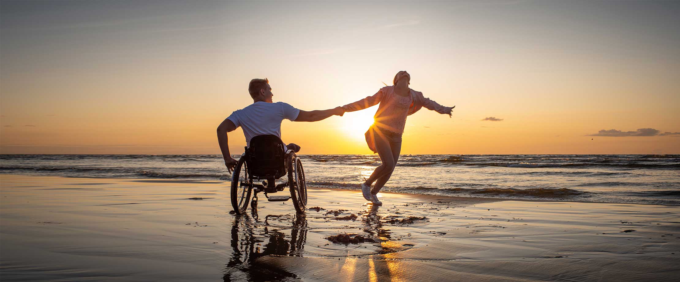 Ein junger Mann im Rollstuhl und eine junge Frau halten sich an den Händen, während sie sich im Kreis drehen. Das paar ist bei Sonnenuntergang am Strand.