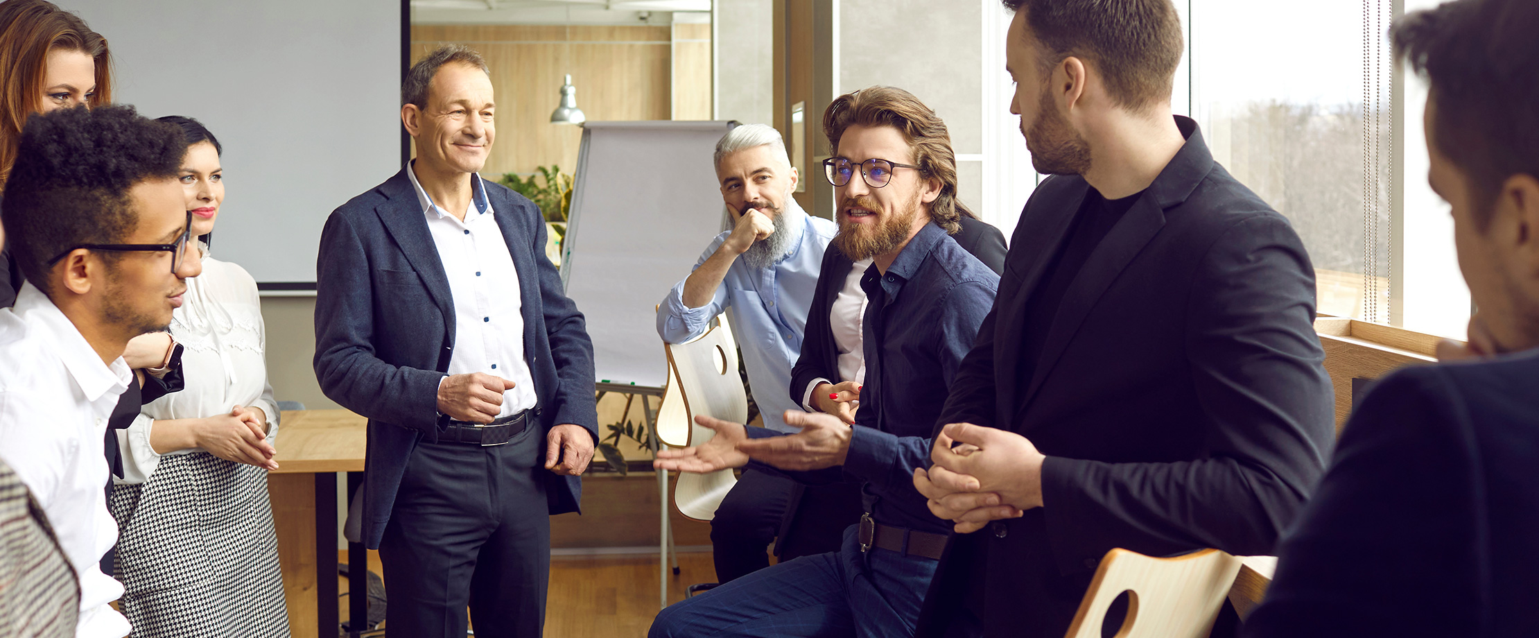 Einige Männer und Frauen in Business-Kleidung stehen und sitzen zusammen und unterhalten sich. Ein Mann mit Brille spricht, die anderen sehen ihn dabei an. 